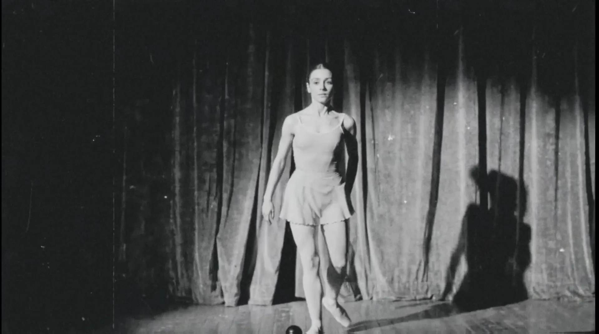 Bailarina sola en un escenario de teatro, de frente bajo un spotlight, su sombra doble proyectada sobre el telón