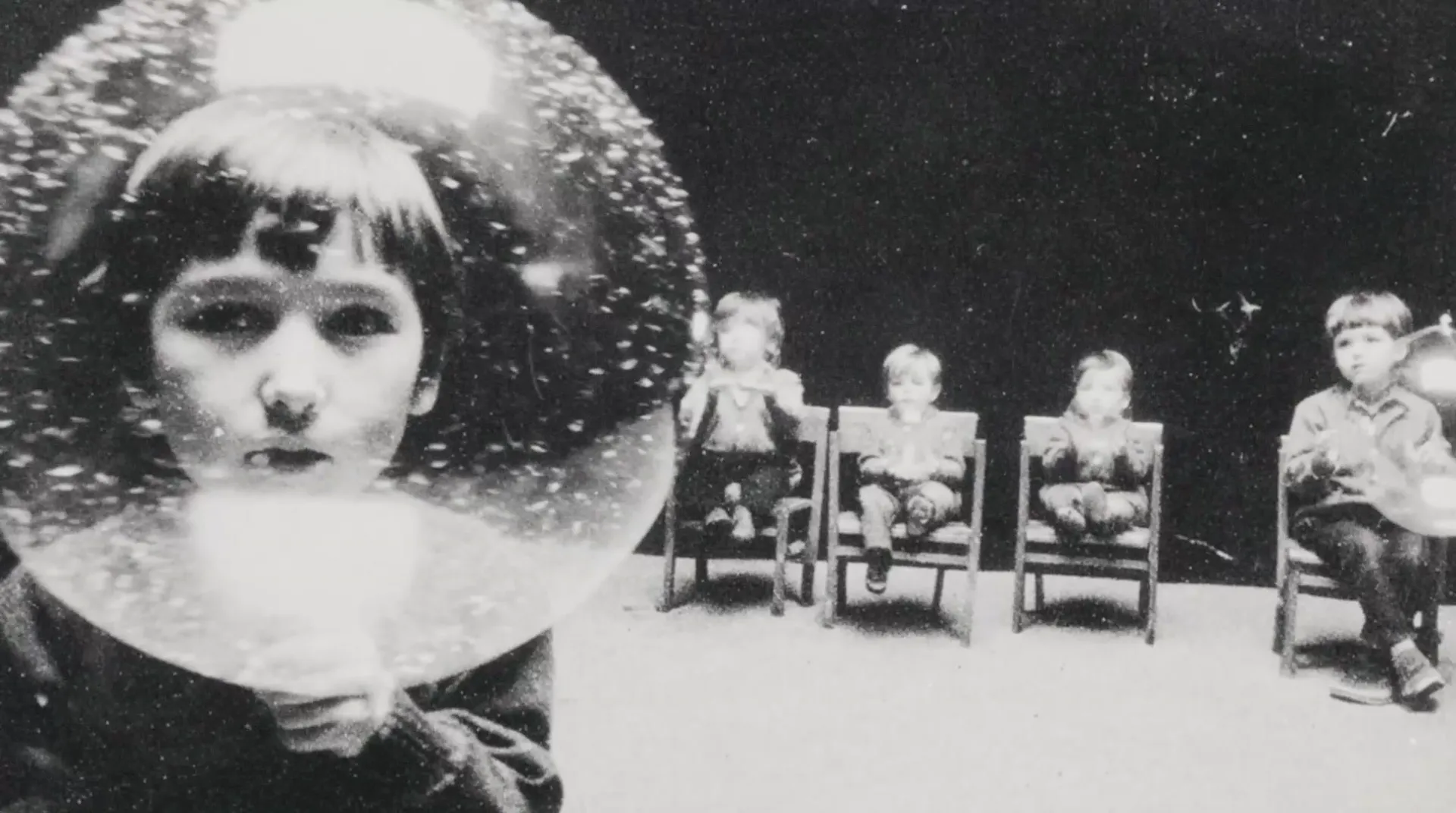 Niña con la cabeza dentro de un globo de vidrio con nieve suspendida, cuatro niños sentados en sillas la observan desde la oscuridad